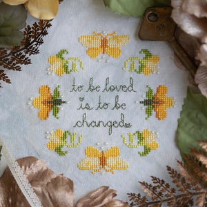 May include: Cross-stitch embroidery on a white fabric with a yellow and green floral design and the text "to be loved is to be changed".