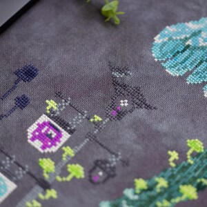 May include: Cross-stitch artwork featuring a nighttime scene with street lamps, a building, and stylized floral elements. The lamps have purple and white accents, and the overall color palette includes shades of gray, purple, green, and blue.