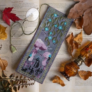 May include: A purple and green cross-stitch bookmark with a mushroom design. The bookmark is on a book with text about role-playing games.