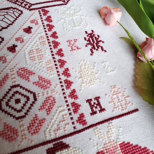 Another view of the finished pink cross stitch sampler of several insects and heart motifs, including a large cicada, praying mantis, and several moths. Focuses on the initials K L H and beetle motif.