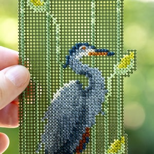 May include: A hand holds a needlepoint canvas featuring a detailed depiction of a blue heron. The bird is rendered in shades of blue, gray, and brown, set against a green background with yellow accents. The canvas is rectangular with a grid of small holes.