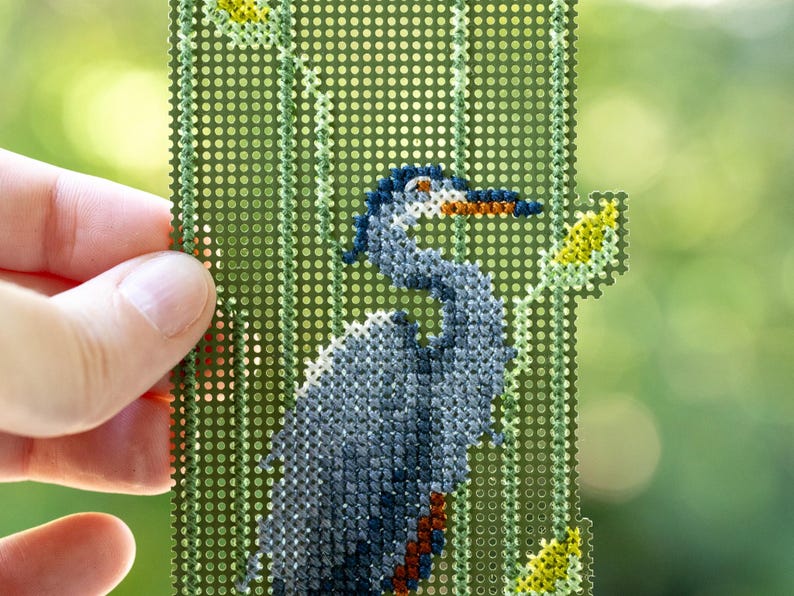 May include: A hand holds a needlepoint canvas featuring a detailed depiction of a blue heron. The bird is rendered in shades of blue, gray, and brown, set against a green background with yellow accents. The canvas is rectangular with a grid of small holes.
