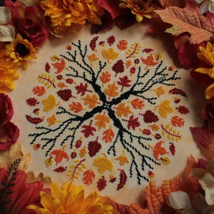 May include: A cross-stitch embroidery design featuring a stylized tree with autumn leaves in shades of red, orange, and yellow. The design is surrounded by a border of fall leaves and flowers.