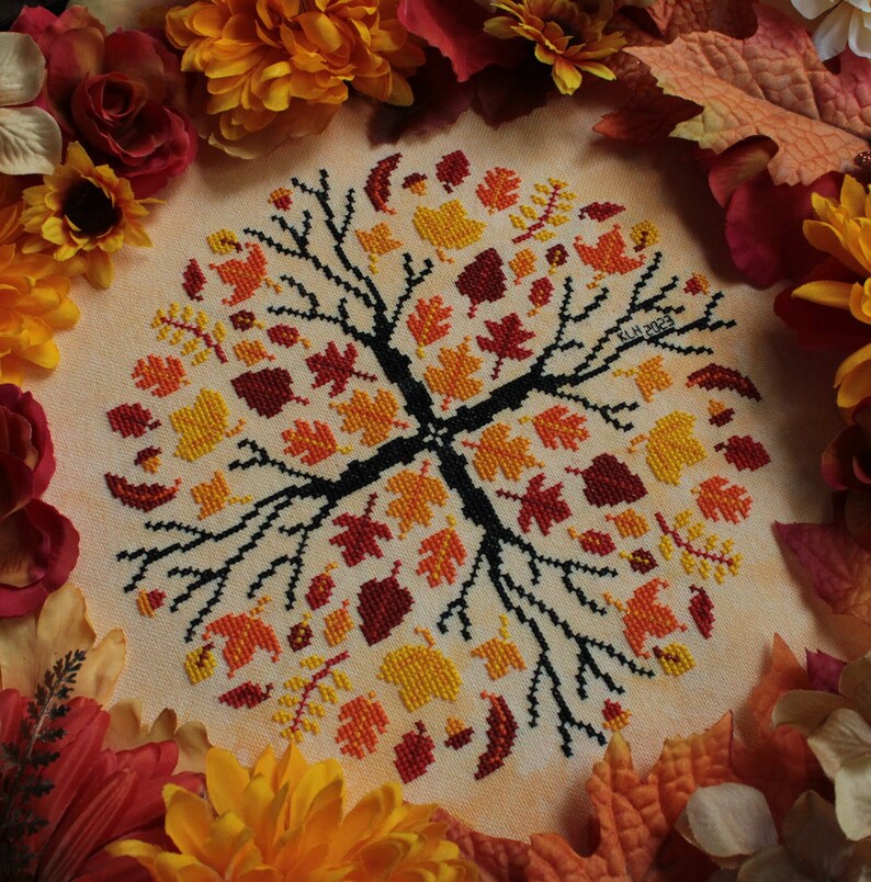 May include: A cross-stitch embroidery design featuring a stylized tree with autumn leaves in shades of red, orange, and yellow. The design is surrounded by a border of fall leaves and flowers.