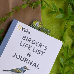 May include: A "Birder's Life List & Journal" book with a bird illustration, a National Geographic logo, and a beaded bookmark. The book is on a brown surface with green foliage and a green felt background.
