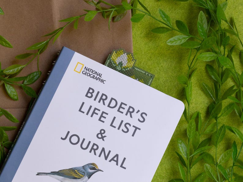 May include: A "Birder's Life List & Journal" book with a bird illustration, a National Geographic logo, and a beaded bookmark. The book is on a brown surface with green foliage and a green felt background.