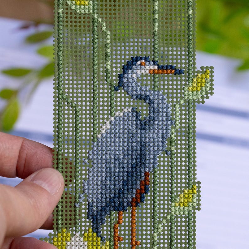 May include: A needlepoint bookmark featuring a blue heron. The bird is stitched against a green background with yellow and green floral accents. The heron has a long orange beak and is standing on orange legs.