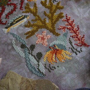 K&ouml;nnte beinhalten: Kreuzstich-Stickerei mit einem maritimen Motiv, das verschiedene Meeresbewohner und Pflanzen zeigt. Das Design beinhaltet einen Fisch, Algen, Korallen und einen Seestern. Der Text "Sea You Soon" ist in der rechten unteren Ecke gestickt.