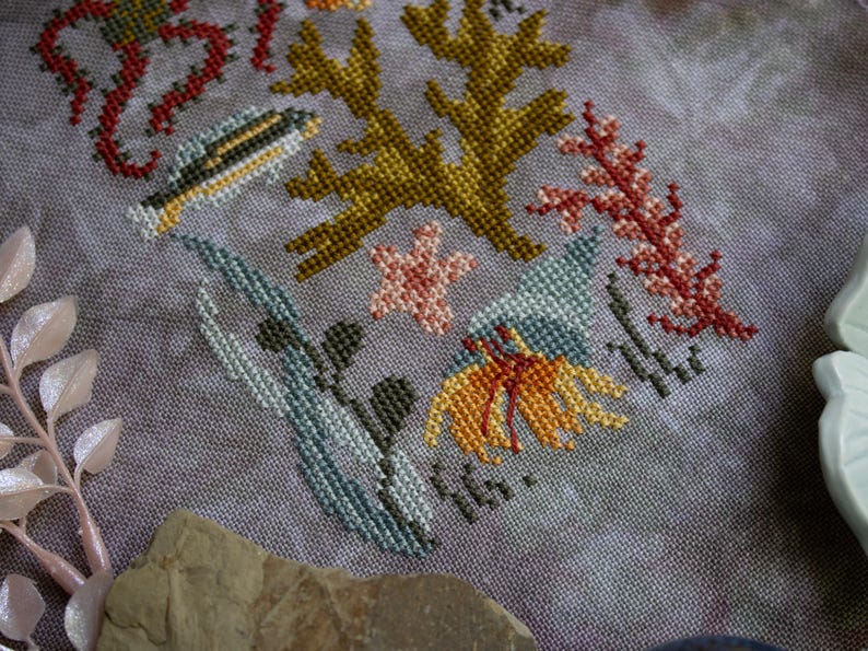 K&ouml;nnte beinhalten: Kreuzstich-Stickerei mit einem maritimen Motiv, das verschiedene Meeresbewohner und Pflanzen zeigt. Das Design beinhaltet einen Fisch, Algen, Korallen und einen Seestern. Der Text "Sea You Soon" ist in der rechten unteren Ecke gestickt.