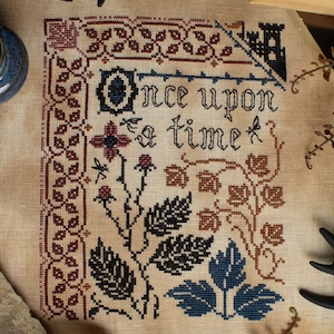 May include: Cross-stitch embroidery on beige fabric with the text "Once upon a time" in black thread. The design features a floral motif with red, brown, and blue leaves.