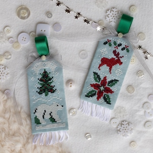 以下が含まれることがあります： 淡い青色の背景に刺繍された2つのクリスマスオーナメント。1つはクリスマスツリーとホッキョクグマ、もう1つは赤いトナカイとポインセチアが特徴です。どちらも緑のリボンループと白いフリンジが付いています。