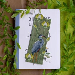May include: A handmade bookmark featuring a detailed beaded design of a blue heron bird and yellow flowers on a green background. The bookmark is placed on a book titled "Birds Life Journal".