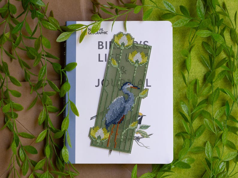 May include: A handmade bookmark featuring a detailed beaded design of a blue heron bird and yellow flowers on a green background. The bookmark is placed on a book titled "Birds Life Journal".