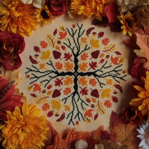 May include: A cross-stitch embroidery of a tree with autumn leaves in shades of red, orange, and yellow. The tree is surrounded by a border of fall leaves and flowers.