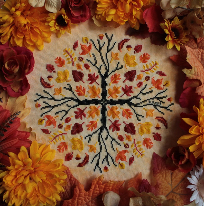 May include: A cross-stitch embroidery of a tree with autumn leaves in shades of red, orange, and yellow. The tree is surrounded by a border of fall leaves and flowers.