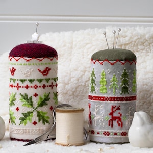 May include: Two cylindrical pincushions with cross-stitch patterns. One has a burgundy top and a design of birds and snowflakes. The other has a green top and a pattern of trees and reindeer. A spool of thread and scissors are also present.