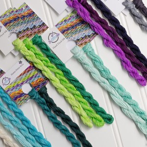 May include: A collection of hand-dyed silk threads in various colors, including shades of blue, green, purple, and gray. Each thread is neatly braided and displayed with a small tag. The tags read "Hand Dyed Silk".