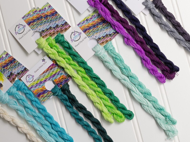 May include: A collection of hand-dyed silk threads in various colors, including shades of blue, green, purple, and gray. Each thread is neatly braided and displayed with a small tag. The tags read "Hand Dyed Silk".