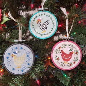 May include: Three handmade Christmas decorations featuring embroidered images of chickens. The decorations are round and have a white background with colourful embroidery. The first decoration features a black and white chicken with a red hat, the second decoration features a yellow chicken with a blue hat, and the third decoration features a red chicken with a green hat.