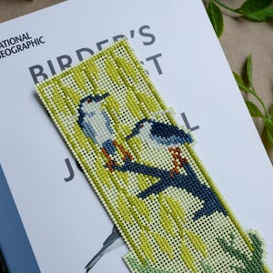 May include: A rectangular cross-stitch bookmark featuring two birds perched on a dark blue branch against a light yellow background with green foliage. The embroidery is worked on a grid-like fabric with visible holes throughout the design.