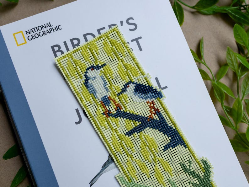 May include: A rectangular cross-stitch bookmark featuring two birds perched on a dark blue branch against a light yellow background with green foliage. The embroidery is worked on a grid-like fabric with visible holes throughout the design.