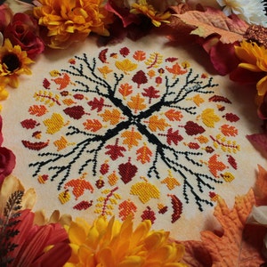May include: A cross-stitch embroidery design featuring a stylized tree with branches and leaves in shades of red, orange, and yellow. The design is surrounded by a border of fall-colored leaves.
