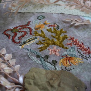 K&ouml;nnte beinhalten: Ein Kreuzstich-Stickmuster mit verschiedenen Meeresbewohnern, darunter Korallen, Algen und Fische. Das Muster ist auf einem hellvioletten Stoff mit einer dezenten Textur gestickt.