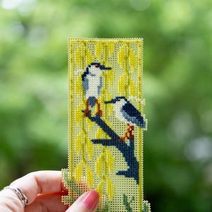 May include: A rectangular cross-stitch bookmark on light green plastic canvas featuring two birds with white and dark blue plumage perched on a dark blue branch, surrounded by yellow willow leaves stitched in a vertical pattern.