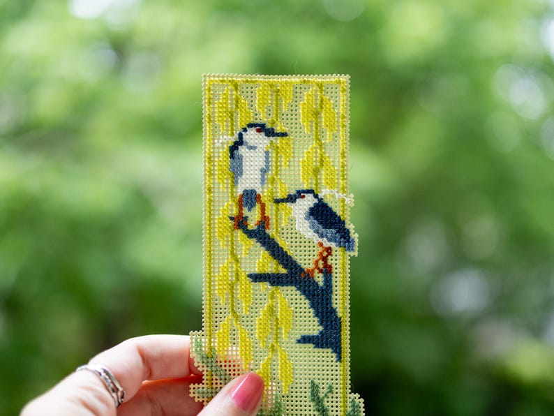May include: A rectangular cross-stitch bookmark on light green plastic canvas featuring two birds with white and dark blue plumage perched on a dark blue branch, surrounded by yellow willow leaves stitched in a vertical pattern.