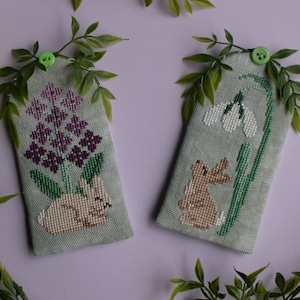 May include: Two cross-stitch needlepoint ornaments featuring a bunny rabbit with a bouquet of purple flowers and a bunny rabbit with a single white snowdrop flower.