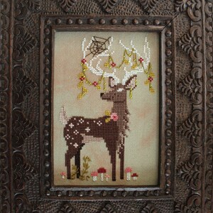 May include: Cross-stitch art of a deer with white antlers adorned with flowers and a spiderweb, set against a neutral background. The deer is brown with white spots and pink flowers. The artwork is framed in a dark, intricately carved wooden frame.