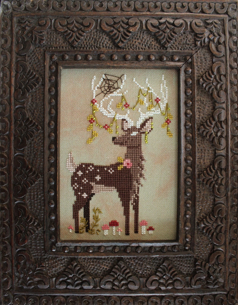 May include: Cross-stitch art of a deer with white antlers adorned with flowers and a spiderweb, set against a neutral background. The deer is brown with white spots and pink flowers. The artwork is framed in a dark, intricately carved wooden frame.