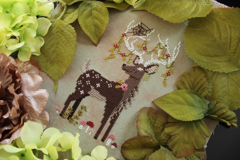May include: A cross-stitch design of a deer with white spots and antlers, adorned with flowers and berries. The deer is surrounded by greenery and mushrooms, creating a woodland theme. The antlers have a spider web design.