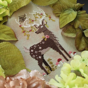 May include: Cross-stitch artwork featuring a brown deer with white spots, adorned with floral accents and a spiderweb. The deer stands amidst small pink and red mushrooms and green foliage. The artwork is framed by green and yellow flowers and leaves.