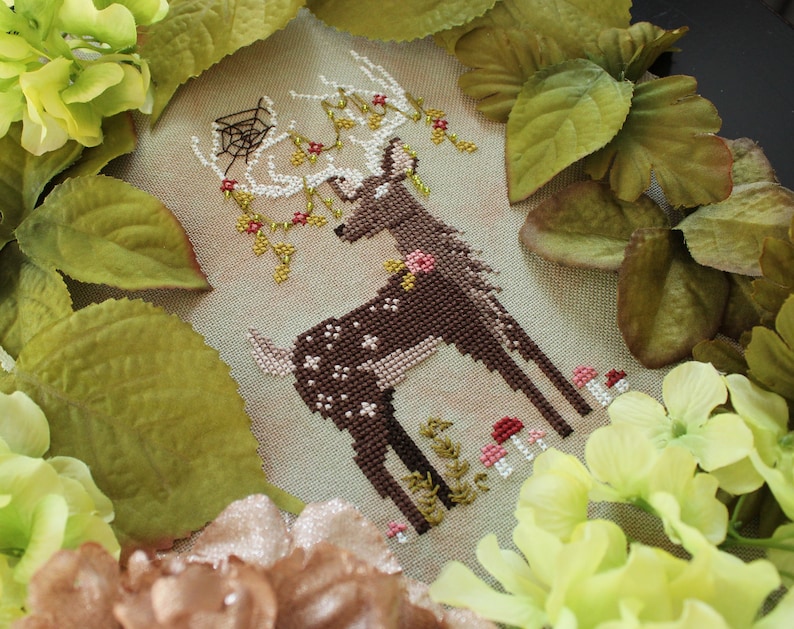 May include: Cross-stitch artwork featuring a brown deer with white spots, adorned with floral accents and a spiderweb. The deer stands amidst small pink and red mushrooms and green foliage. The artwork is framed by green and yellow flowers and leaves.