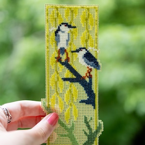 May include: A rectangular plastic canvas bookmark featuring a cross-stitch design of two birds perched on a dark blue branch. The background consists of yellow willow-like foliage and green stems, all stitched onto a light yellow grid base.