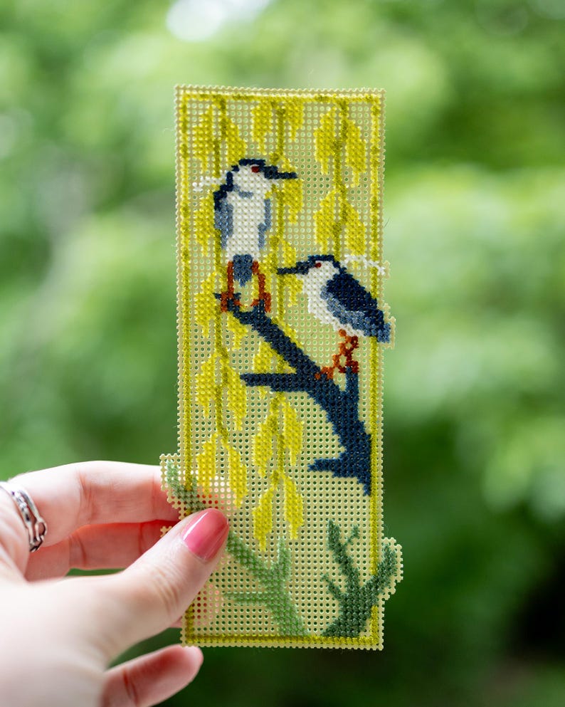 May include: A rectangular plastic canvas bookmark featuring a cross-stitch design of two birds perched on a dark blue branch. The background consists of yellow willow-like foliage and green stems, all stitched onto a light yellow grid base.