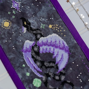 Peut inclure: Une broderie au point de croix détaillée sur le thème céleste. L'œuvre présente un serpent ailé noir et violet sur un fond sombre et étoilé. Le motif comprend une bordure violette et des accents de jaune, de vert et de blanc, avec un collier en argent en forme d'étoile.