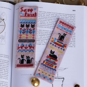 以下が含まれることがあります： 薄紫色の背景に、2つの刺繍されたブックマーク。1つのブックマークには「Save Point」と書かれており、黒猫と幾何学模様が描かれています。もう1つのブックマークには、本の上に黒猫がいます。どちらも開いた本の上にあります。