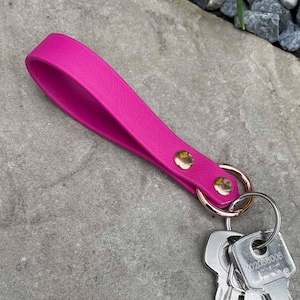 May include: A vibrant pink leather key fob with a loop handle, attached to a rose gold ring with two gold rivets. Several keys are attached. The fob rests on a textured grey surface.