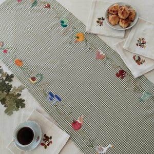 May include: A green and white gingham table runner decorated with embroidered motifs of pumpkins, mushrooms, a snail, a squirrel, and a goose. The set includes matching cream-coloured napkins with similar embroidery and a small plate of biscuits.