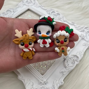 Puede incluir: Tres figuras navideñas de arcilla: un reno con una bufanda roja y blanca, un muñeco de nieve con sombrero negro y bufanda verde, y un hombre de jengibre con un lazo rojo. Las figuras están en una mano, con un marco decorativo blanco en el fondo.