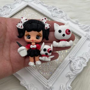 May include: A set of three polymer clay charms featuring a girl with black hair and red bows, a white dog with a red eye, and a white dog with a red eye. The charms are all about 2 inches tall.