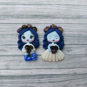 May include: Two handmade polymer clay figurines of a blue-skinned bride with dark blue curly hair, holding a bouquet. One wears a white dress, the other a blue outfit. Both have a floral crown and are on a gray wooden surface.