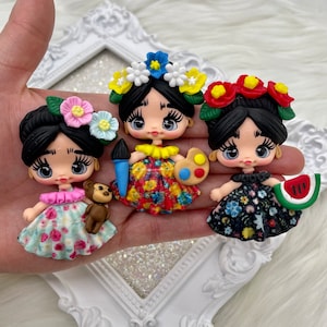 May include: Three colorful handmade polymer clay dolls with black hair, wearing floral dresses and flower crowns. The dolls are holding a paint palette, a paintbrush, and a watermelon slice.