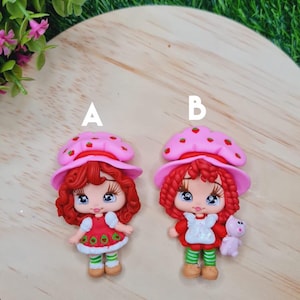 Może przedstawiać: Dwie czerwono-białe figurki lalek Strawberry Shortcake z różowymi kapeluszami i białymi ozdobami. Lalka po lewej trzyma zielono-czerwoną truskawkę. Lalka po prawej trzyma różowego pluszowego misia. Lalki znajdują się na drewnianej powierzchni.