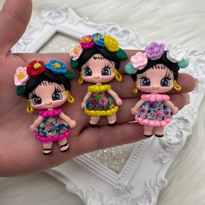 May include: Three handmade doll figurines. Each doll has black hair, a floral crown, and a colorful floral dress. The dolls have gold hoop earrings and are held in a hand.