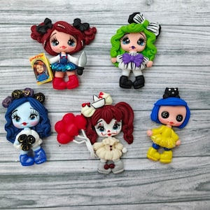Peut inclure: Collection de figurines faites à la main en pâte polymère, chacune représentant une poupée stylisée avec des caractéristiques uniques. Les poupées ont différentes couleurs de cheveux, tenues et accessoires, dont un couteau, un livre et des ballons. Les figurines sont posées sur un fond en bois gris clair.