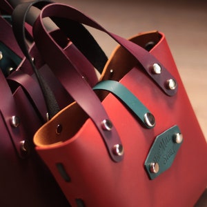 Puede incluir: Primer plano de varios bolsos tote de cuero pequeños en varios colores, incluyendo burdeos, verde azulado y negro. Cada bolso tiene un asa a juego y está asegurado con remaches plateados. Los bolsos tienen forma rectangular y están forrados con un color contrastante.