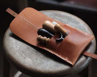 May include: A brown leather pouch with a flap closure and a strap. The pouch contains four tools: a black leather tool, a wooden tool with a white tip, a wooden tool with a brown tip, and a metal tool with a curved blade.